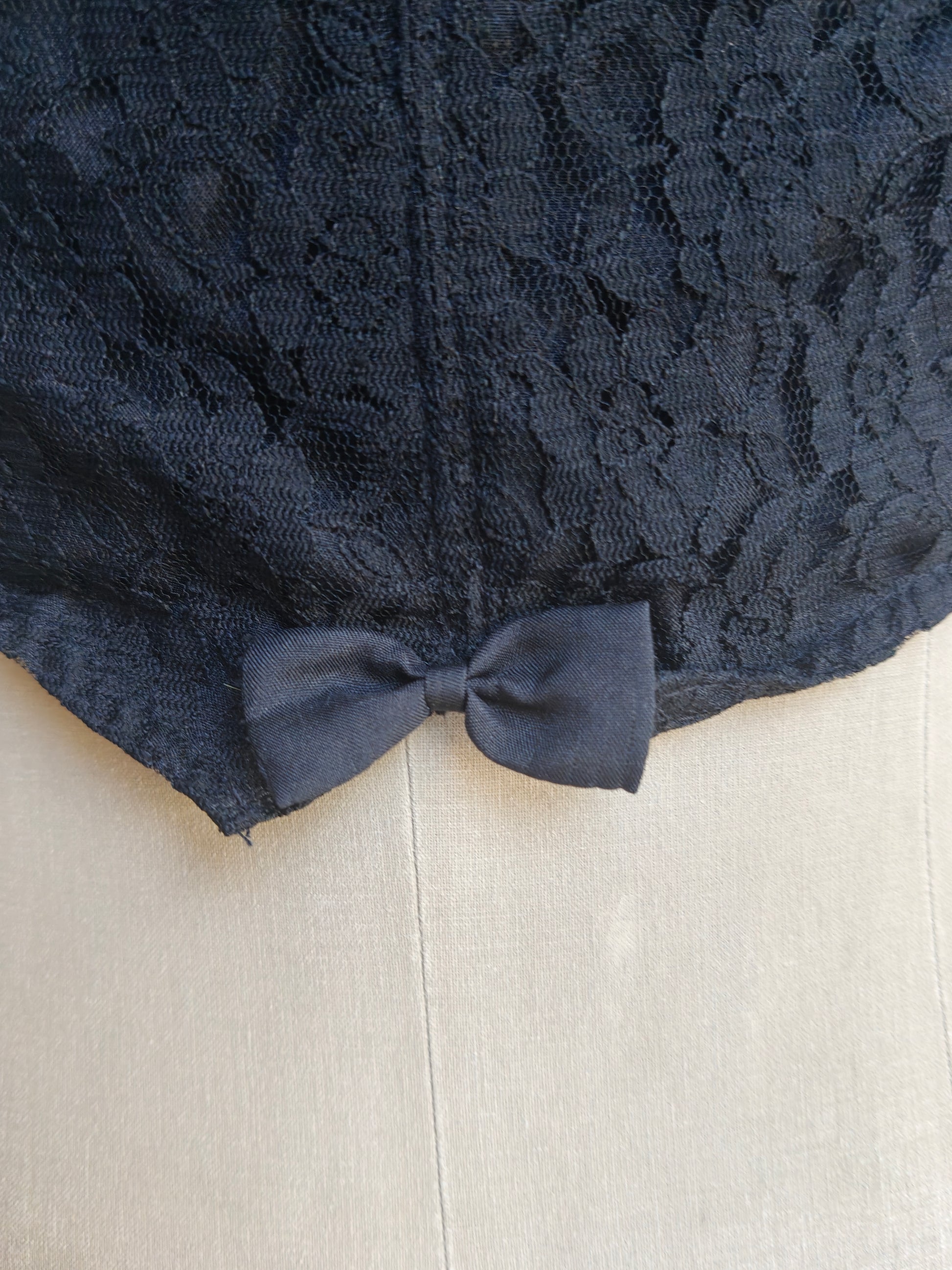 Close-up of the black lace back of top with a matching fabric bow at the hem, displayed on a light beige mannequin or dress form.