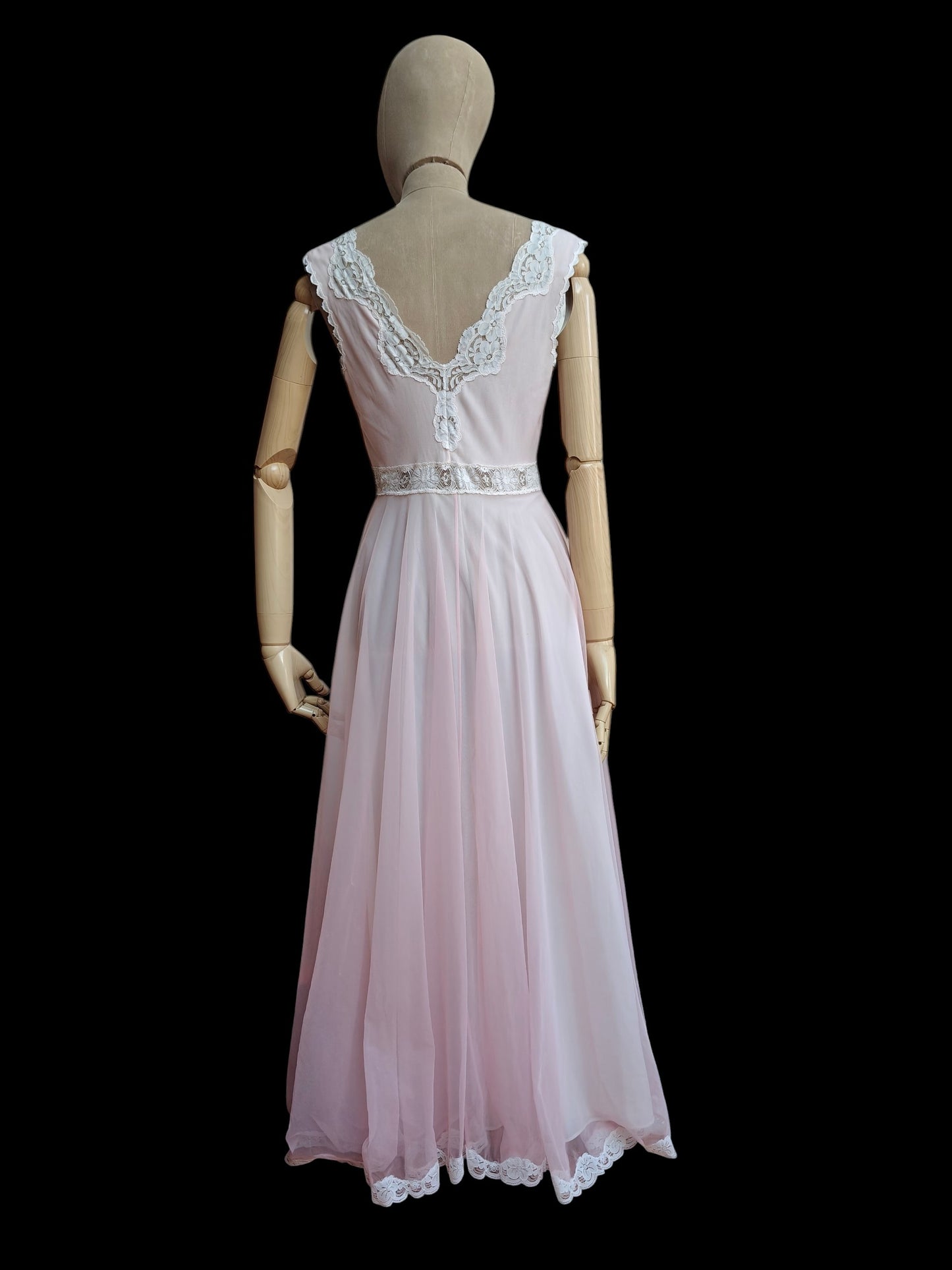 A light pink sleeveless maxi dress displayed on a wooden mannequin from the back, featuring a lace-trimmed V-neckline and a matching lace hem, set against a solid black background.