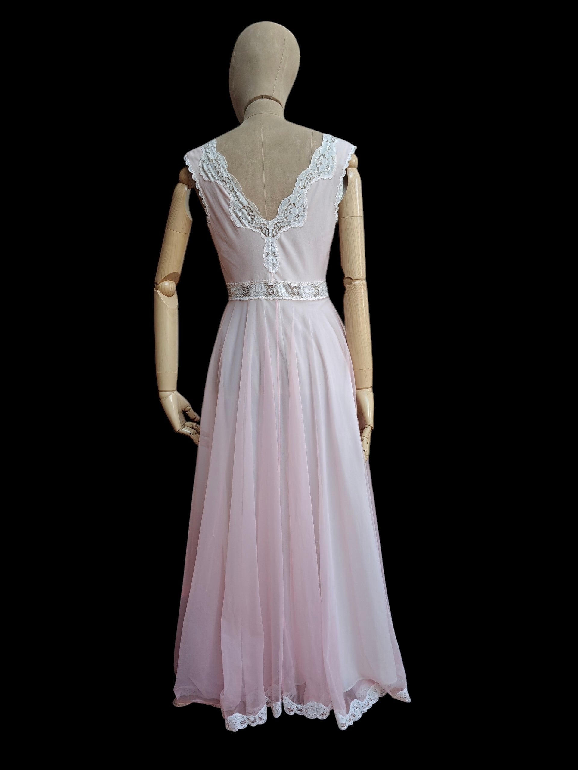 A light pink sleeveless maxi dress displayed on a wooden mannequin from the back, featuring a lace-trimmed V-neckline and a matching lace hem, set against a solid black background.