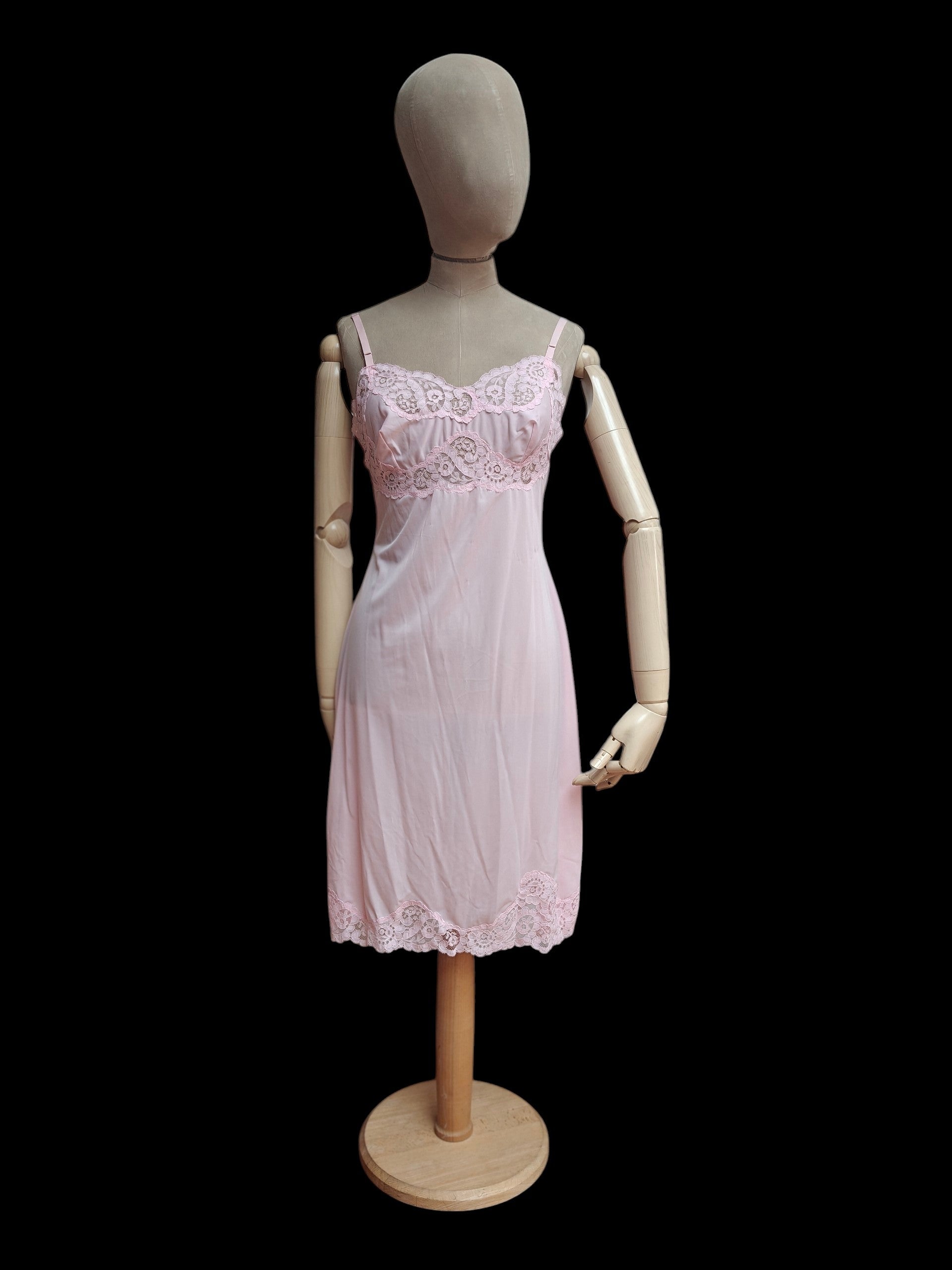 A light pink sheer lingerie chemise with lace trim, displayed on a wooden mannequin against a solid black background.