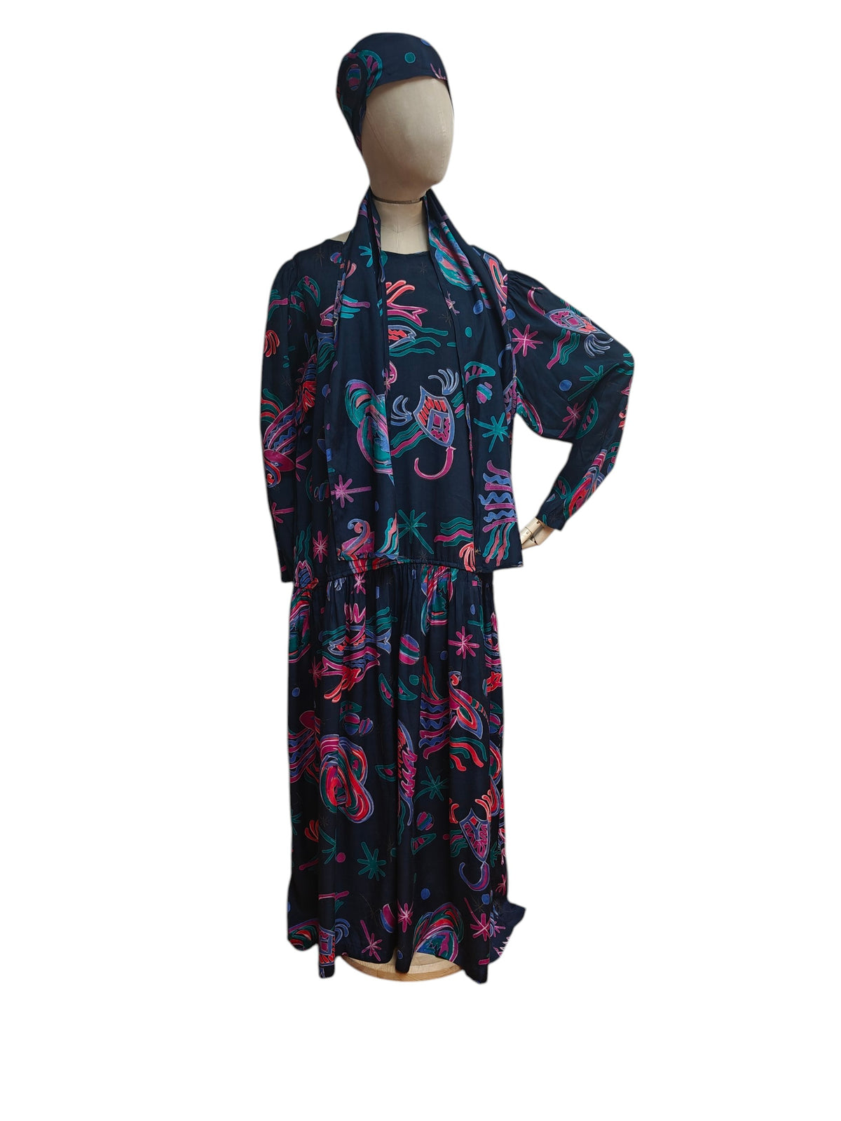 A long-sleeved maxi dress with a matching headscarf displayed on a mannequin. The dress features a dark navy blue base with a vibrant paisley and floral pattern in pink, teal, and purple.