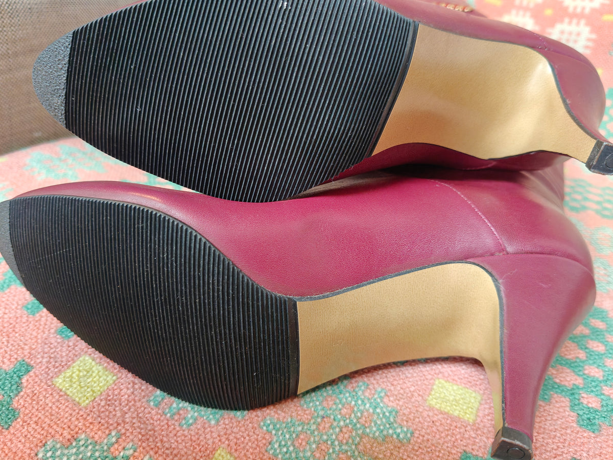 A pair of women's high-heeled pumps shown from the sole, featuring a magenta pink leather upper, a tan insole, and a black ribbed rubber outsole, resting on a patterned pink and green fabric surface.