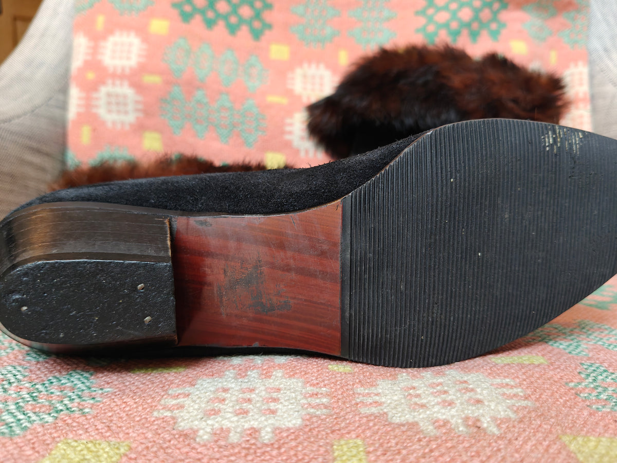 Close-up of the sole of a black suede boot with a reddish-brown wood heel accent and dark brown faux fur trim, resting on a patterned pink and green textile.