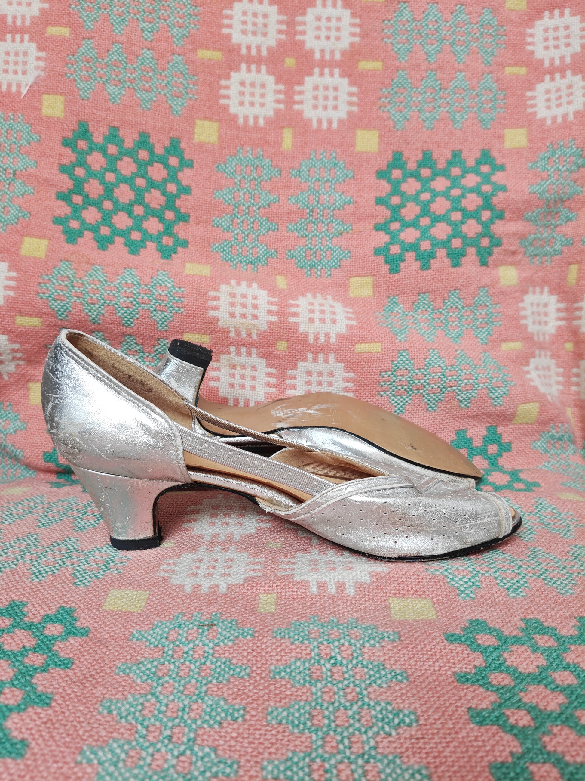 A pair of metallic silver low-heeled pumps with slingback straps, displayed on a pink and green patterned textile background.