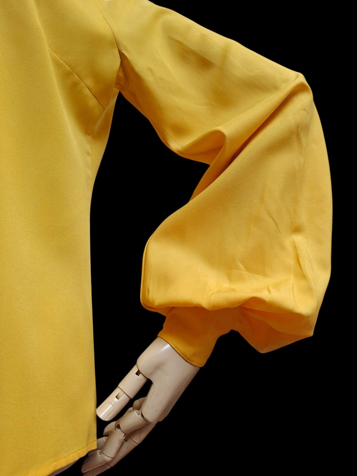 A close-up of a bright yellow long-sleeve garment with a voluminous balloon sleeve, displayed on a mannequin arm against a solid black background.