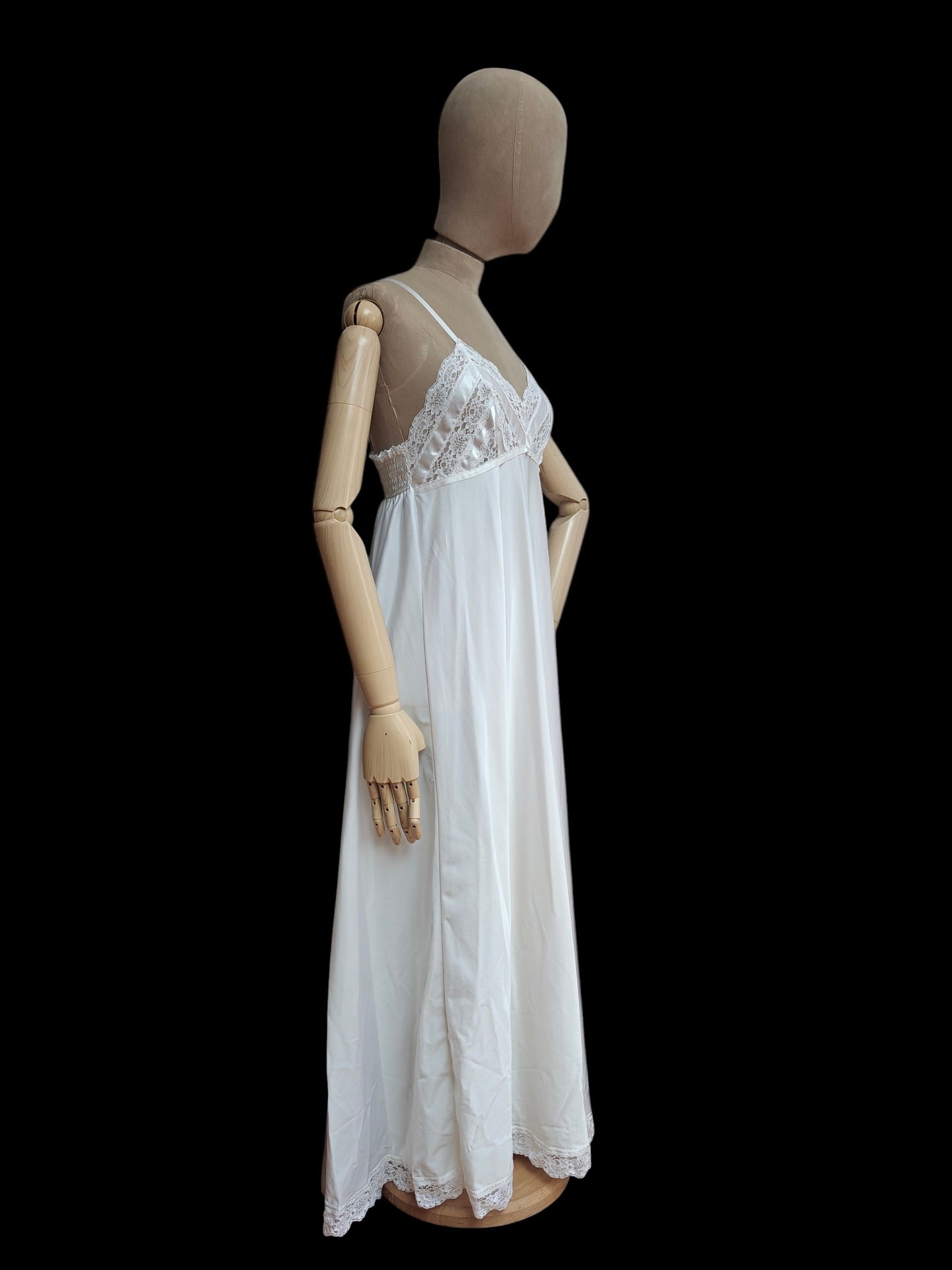 A white slip nightgown with a lace-trimmed bodice and spaghetti straps, displayed on a wooden mannequin against a solid black background.