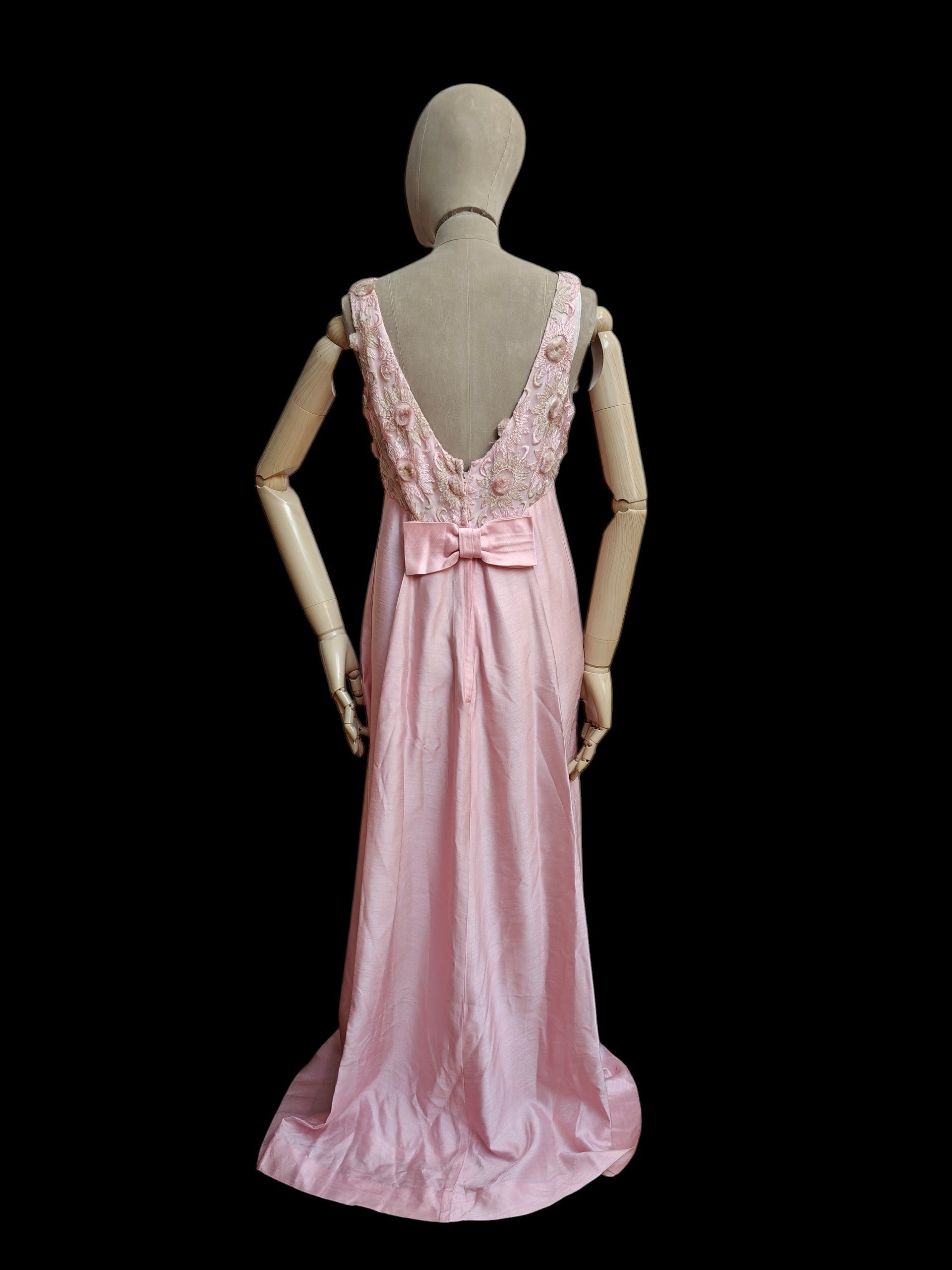 A pink vintage evening gown displayed on a beige mannequin from the back, featuring a deep V-neckline with floral lace applique and a large bow at the waist, set against a solid black background.