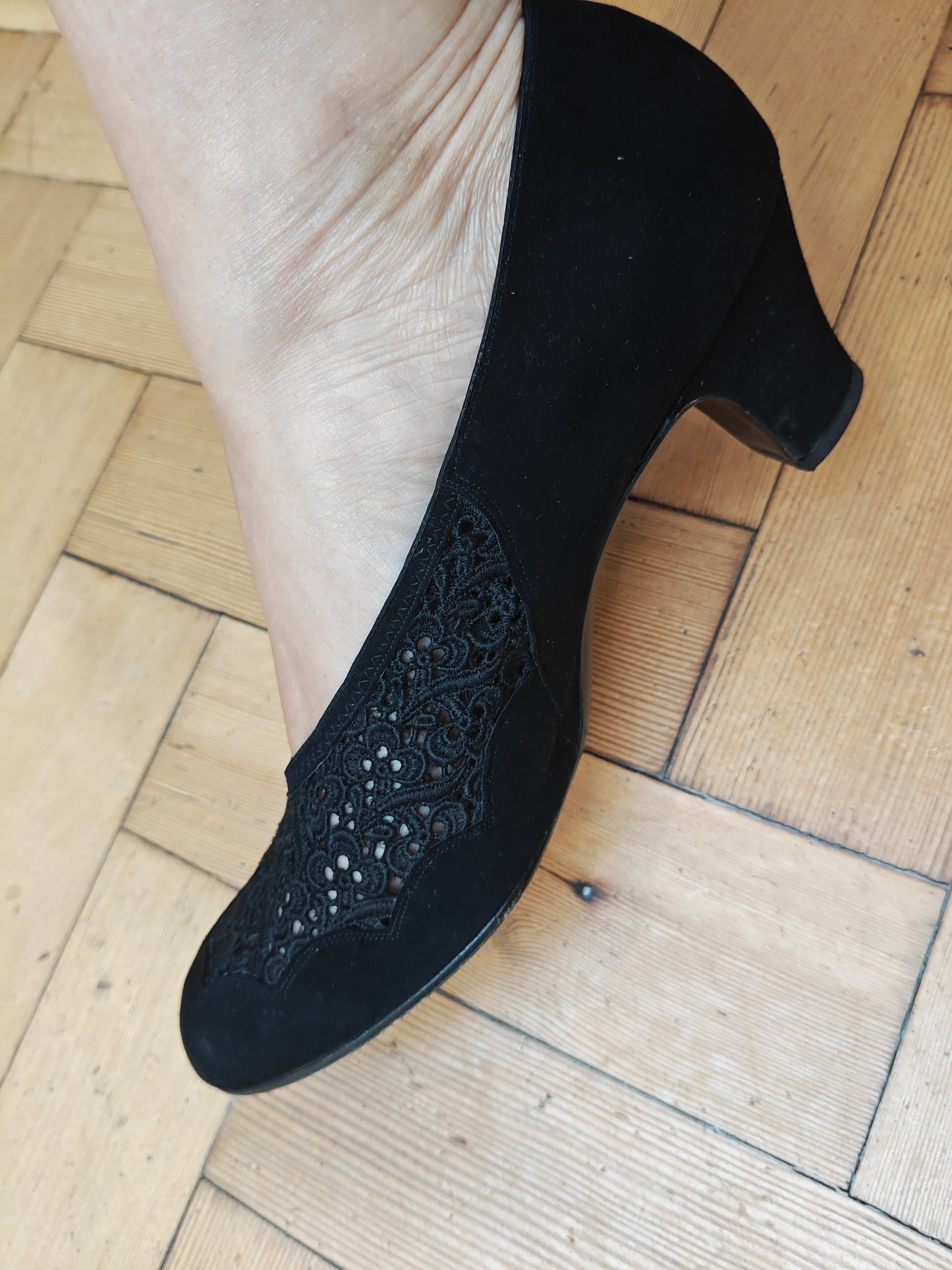 A black suede high-heeled shoe with an intricate floral lace overlay on the side, shown being worn on a foot over a light-colored wooden parquet floor.