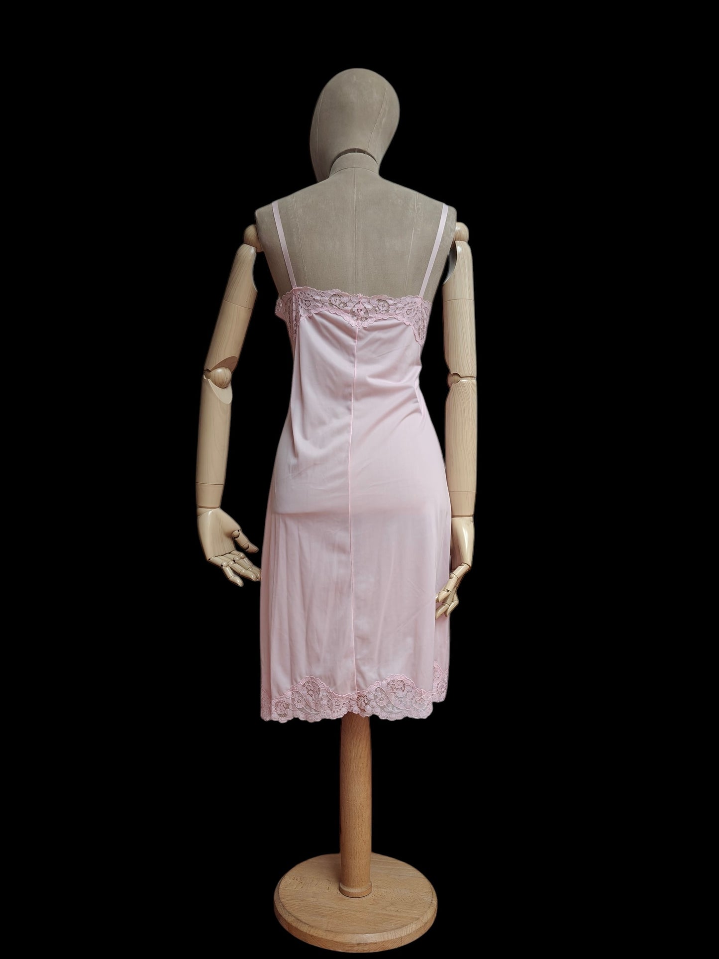 A light pink vintage slip dress with lace trim, displayed on a wooden mannequin from the back against a solid black background.