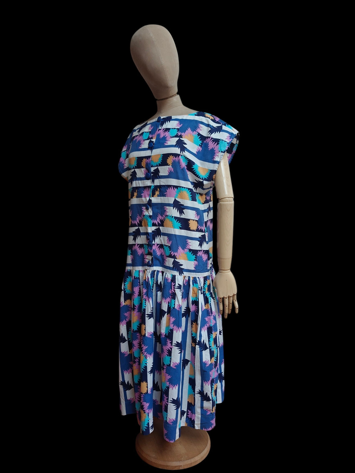 A short-sleeve vintage dress displayed on a mannequin against a black background. The dress features a colorful abstract pattern with blue, pink, purple, yellow, and white shapes.