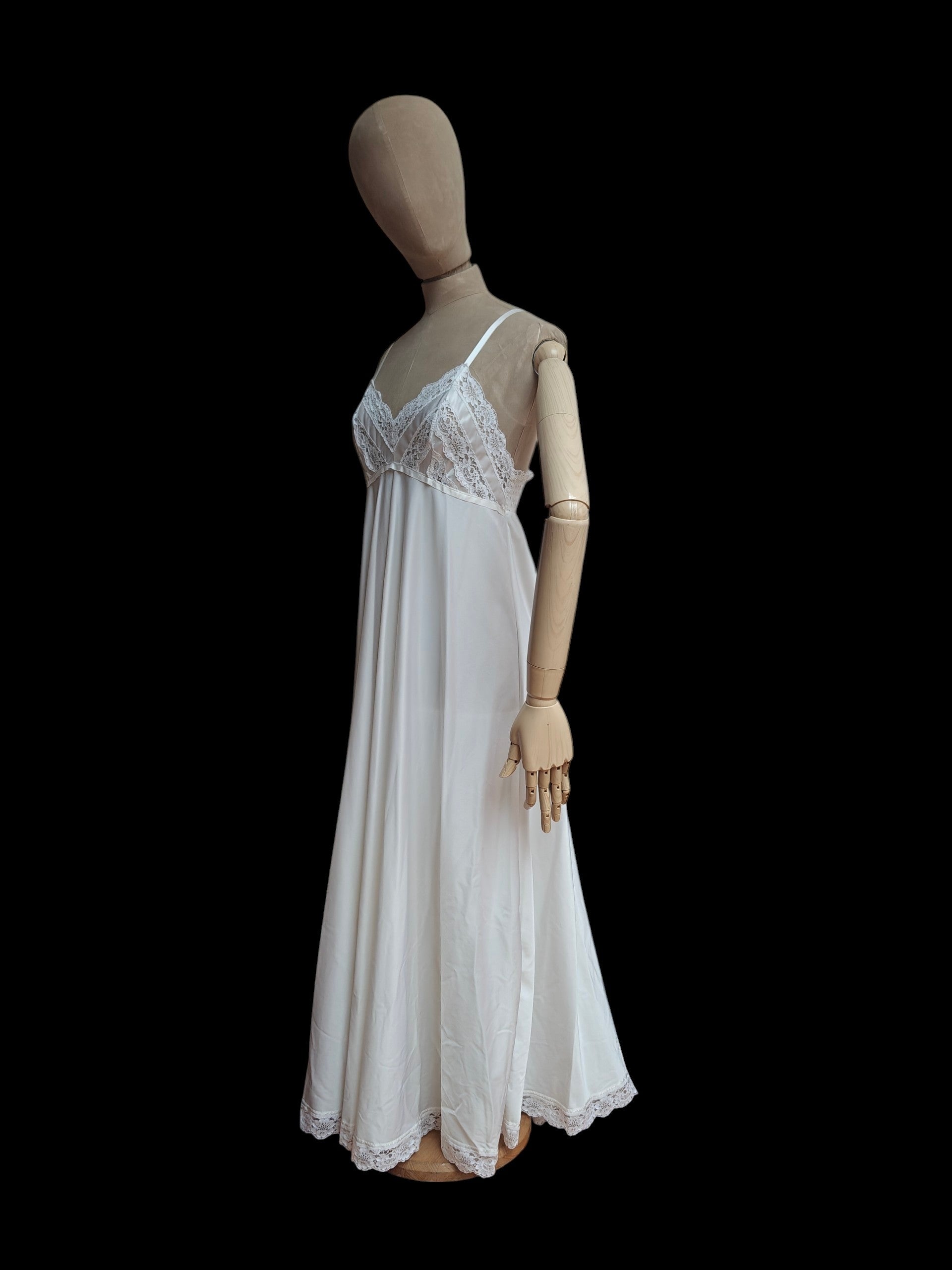 A white vintage nightgown with lace cups and a flowing skirt, displayed on a wooden mannequin against a solid black background.